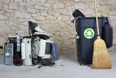 Old electronics sitting next to a recycling bin and broom.