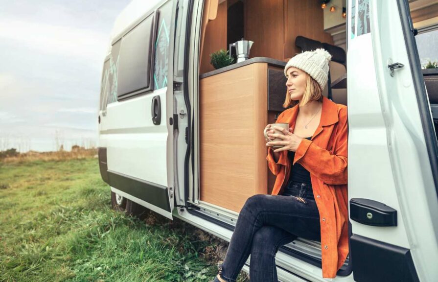 A woman sits in the open door of her camper van enjoying coffee and a view