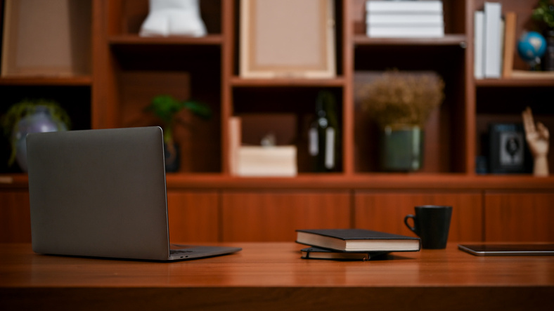 A laptop, mug, and pile of journals sit on a wooden table with shelves in the background.