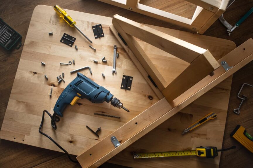 A power drill, screws, and other tools sit on top of a wooden furniture piece that is being assembled.